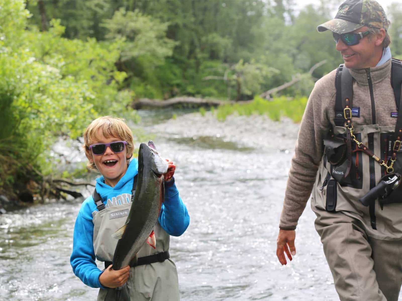 Father and son fishing in an Alaskan river with the son holding a fish they caught.